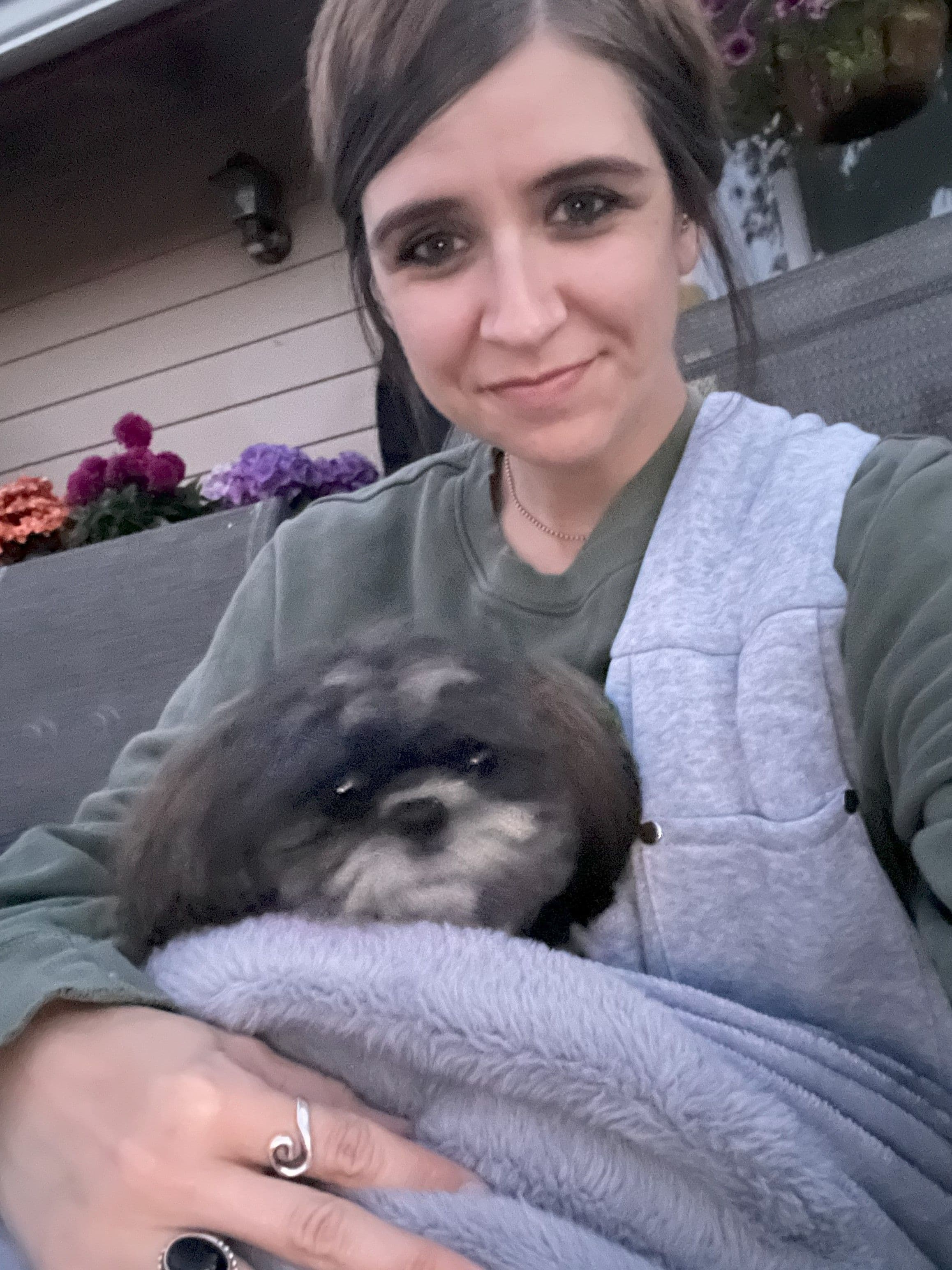 Woman holding a small dog wrapped in a blanket, sitting outdoors with flowers in the background.