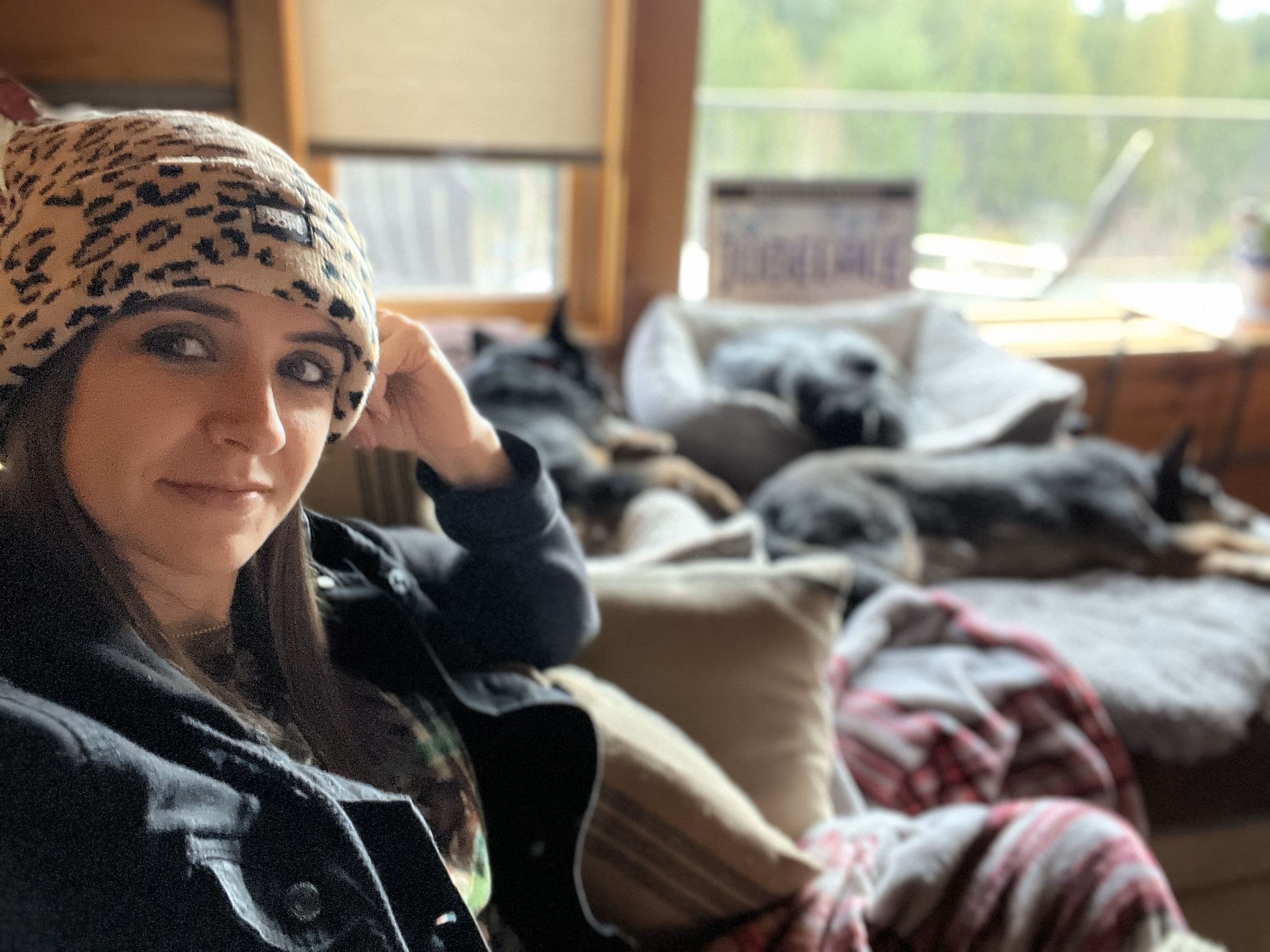 Person in a leopard print beanie relaxing indoors with sleeping dogs on cozy blankets.