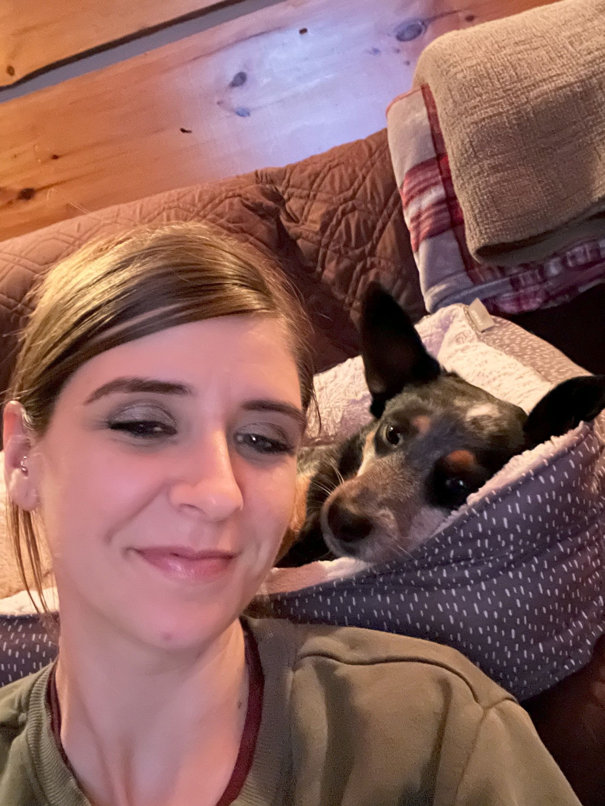 Woman smiles for a selfie with her dog nestled in a cozy bed on a couch.